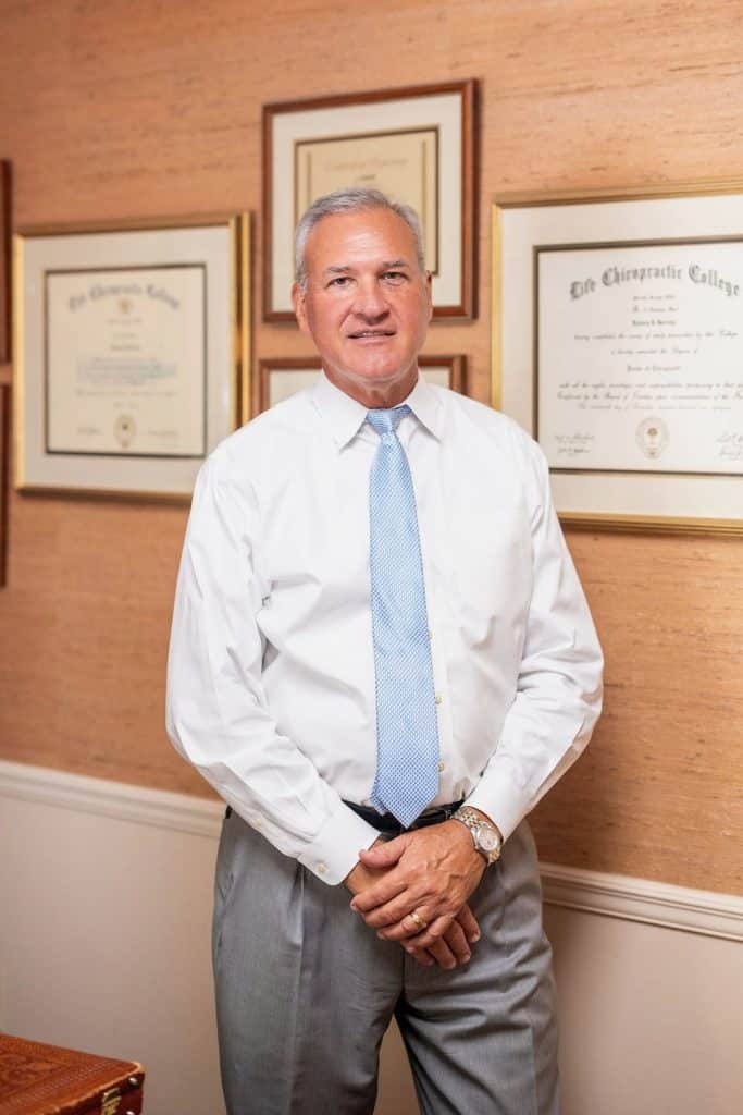 Chiropractor in Opelika, AL office, standing with framed diplomas on wall. Expert care for back pain and wellness.